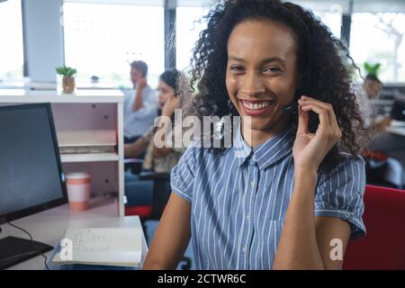Porträt einer smarten Geschäftsfrau mit gemischtem Rennen, die am Schreibtisch sitzt und ein Telefonkopfhörer trägt und vor der Kamera lächelt, mit Kollegen im Hintergrund. Kreative Arbeit Stockfoto