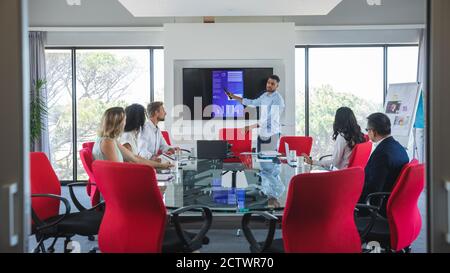 Gemischter Rennunternehmer, der neben einem Bildschirm steht und den männlichen und weiblichen Kollegen, die in einem Besprechungsraum sitzen, eine Präsentation gibt. Kreativer Business-Profi Stockfoto