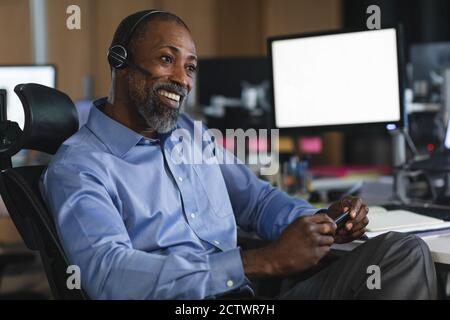 Afroamerikanischer Geschäftsmann, der spät abends in einem modernen Büro arbeitet, an einem Schreibtisch sitzt, ein Headset trägt und lächelt. Stockfoto