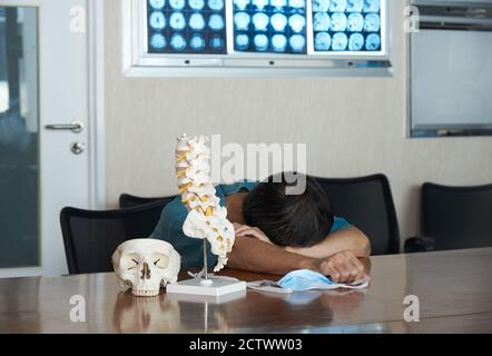 Ein asiatischer Neurochirurg fühlt sich müde und schläft in der medizinischen Praxis. Schädel- und Lendenwirbelsäule Modell auf dem Tisch, Gehirn-MRT an Bord. Stockfoto