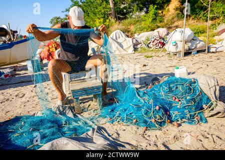 Ein Fischer sitzt auf einem Stuhl am Strand, stapelt Fischernetz auf und reinigt es von Muscheln. Stockfoto