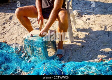 Ein Fischer sitzt auf einem Stuhl am Strand und reinigt Fischernetz von Muscheln, zerschlagen sie mit einem Holzbrett. Stockfoto