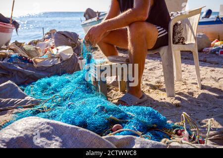 Ein Fischer sitzt auf einem Stuhl am Strand, stapelt Fischernetz auf und reinigt es von Muscheln. Stockfoto