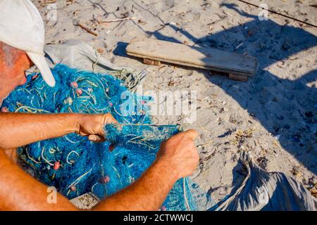 Ein Fischer sitzt auf einem Stuhl am Strand, stapelt Fischernetz auf und reinigt es von Muscheln. Stockfoto