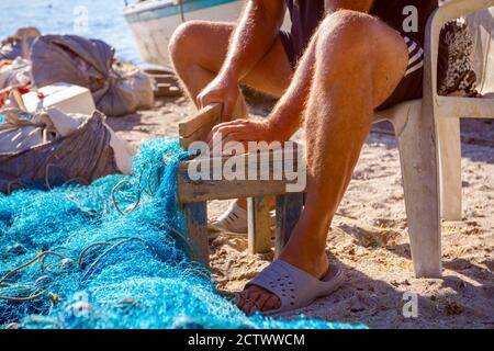 Ein Fischer sitzt auf einem Stuhl am Strand und reinigt Fischernetz von Muscheln, zerschlagen sie mit einem Holzbrett. Stockfoto