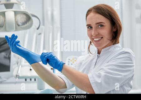 Eine Zahnärztin zieht Handschuhe vor dem Hintergrund der zahnärztlichen Ausrüstung in einer Zahnarztpraxis an. Glückliches Patienten- und Zahnarztkonzept. Stockfoto