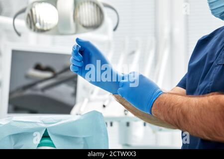 Der männliche Zahnarzt zieht Handschuhe vor dem Hintergrund der zahnärztlichen Ausrüstung in einer Zahnarztpraxis an. Glückliches Patienten- und Zahnarztkonzept.