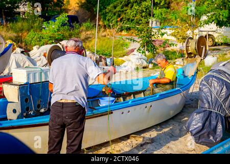 Makrygialos, Griechenland - 29. August 2018: Zwei Fischer am Strand, einer im Boot und der andere neben dem Boot, stapeln Fischnetz für das nächste Angeln. Stockfoto