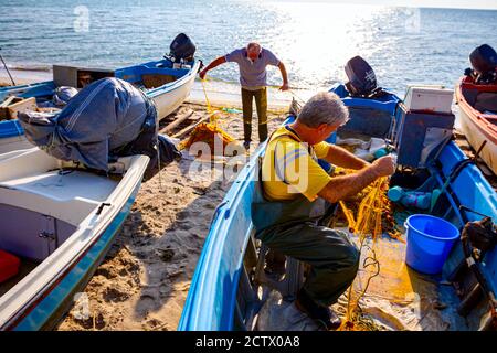 Zwei Fischer am Strand, einer im Boot und der andere neben dem Boot, stapeln Fischnetz für das nächste Angeln. Stockfoto