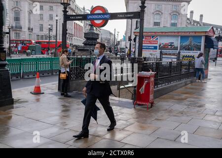 Nach einem Regenschauer im Zentrum Londons - dem sechsmonatigen Punkt der Coronaviruus Pandemie-Sperre - schreitet ein Geschäftsmann mit Anzug und Gesichtsbedeckung (über dem Mund, aber nicht über der Nase) am 24. September in London, England, durch den Piccadilly Circus mit einem Stechbauch. Nach einem plötzlichen Anstieg der Covid-Infektionsrate, einem vorhergesagten „zweiten Anstieg“, werden von der Regierung wieder neue Beschränkungen eingeführt. Und nachdem sie zurück ins Büro ermutigt wurden, um den lokalen Volkswirtschaften zu helfen, werden die Arbeitnehmer wieder aufgefordert, wenn möglich von zu Hause aus zu arbeiten. Stockfoto