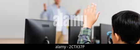 Horizontale Ernte der asiatischen Schuljunge mit erhobener Hand sitzt in der Nähe Computer in der MINT-Schule Stockfoto