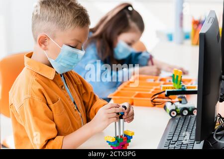 Selektiver Fokus der Schuljunge in der medizinischen Maske Modellierung Roboter aus Bausteine und Computer in der Schule Stockfoto