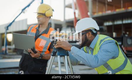 Auf der Baustelle für gewerbliche Industriegebäude: Der professionelle Ingenieur Surveyor ergreift Maßnahmen mit Theodolite, Worker nutzt Laptop. Im Stockfoto