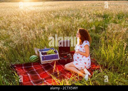 Eine schwangere Frau sitzt an einem sonnigen Tag auf der roten Decke neben einem Grammophon und einem Korb mit Früchten auf dem Weizenfeld. Stockfoto