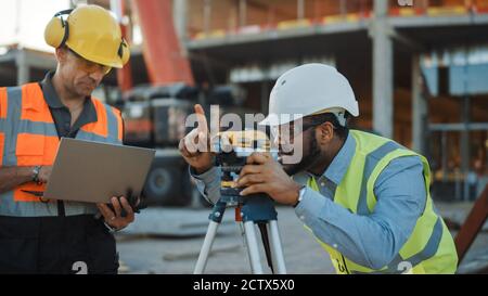 Auf der Baustelle für gewerbliche Industriegebäude: Der professionelle Ingenieur Surveyor ergreift Maßnahmen mit Theodolite, Worker nutzt Laptop. Im Stockfoto