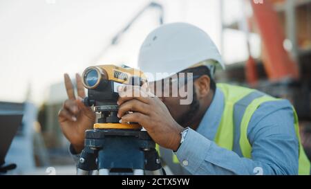 Auf der Baustelle für gewerbliche Industriegebäude: Der professionelle Ingenieur Surveyor ergreift Maßnahmen mit Theodolite, Worker nutzt Laptop. Im Stockfoto