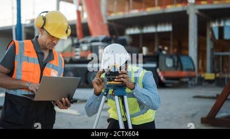 Im Inneren der kommerziellen Industriebaustelle: Der professionelle Ingenieur Surveyor nimmt mit Theodolite Maßnahmen mit Hilfe von Digital Tablet Stockfoto