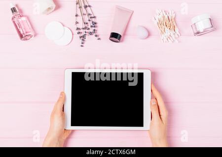 Hände der Frau halten elektronische Tablette mit leerem Bildschirm neben kosmetischen Cremes, Lotionen, Lavendel-Aromaöl und Hygienezubehör auf rosa Tisch, Stockfoto