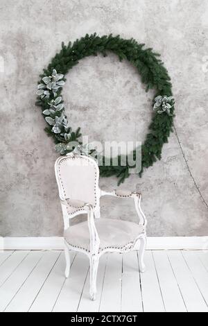 Weihnachtliche Innenausstattung des Zimmers. Grauer Stuhl steht unter einer hellen Wand, an der ein Weihnachtskranz hängt. Grüner Nadelkranz mit Silber verziert Stockfoto