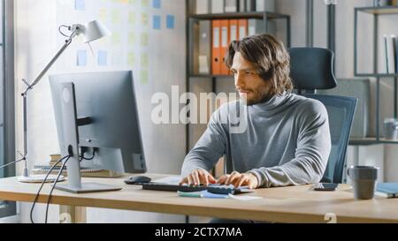 Schöner langhaariger Unternehmer sitzt an seinem Schreibtisch im Büro arbeitet auf Desktop-Computer, arbeiten mit Dokumenten, Charts, Grafiken, Statistik und Stockfoto