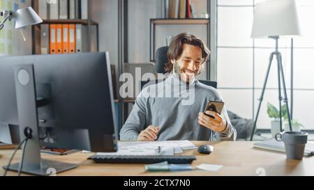 Schöner langhaariger Unternehmer sitzt an seinem Schreibtisch im Büro arbeitet auf Desktop-Computer, Arbeiten mit Dokumenten, Grafiken. Lächelnd Verwendet Stockfoto