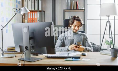 Schöner langhaariger Unternehmer sitzt an seinem Schreibtisch im Büro arbeitet auf Desktop-Computer, Arbeiten mit Dokumenten, Grafiken. Verwendet Smartphone, Social Media Stockfoto