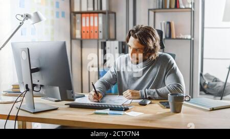 Gutaussehende langhaarige Unternehmer sitzen an seinem Schreibtisch im Büro arbeitet auf Desktop-Computer, Arbeiten mit Dokumenten, Korrigieren von Diagrammen, Zeichnen Stockfoto