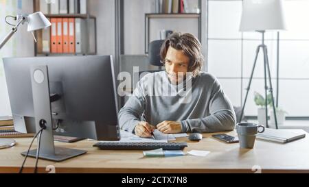 Gutaussehende langhaarige Unternehmer sitzen an seinem Schreibtisch im Büro arbeitet auf Desktop-Computer, Arbeiten mit Dokumenten, Korrigieren von Diagrammen, Zeichnen Stockfoto