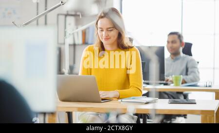 Schöne und kreative junge Frau sitzen an ihrem Schreibtisch mit Laptop-Computer. Im Hintergrund Bright Office wo Diverse Team von Young Stockfoto