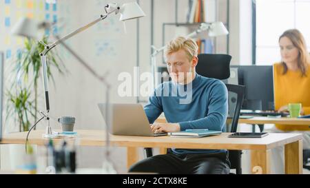 Der gutaussehende kaukasische IT-Spezialist, der an seinem Schreibtisch sitzt, arbeitet im Rahmen seines Geschäftsprojekts auf einem Laptop. Vielfältiges Team von Young Professionals, in denen gearbeitet wird Stockfoto