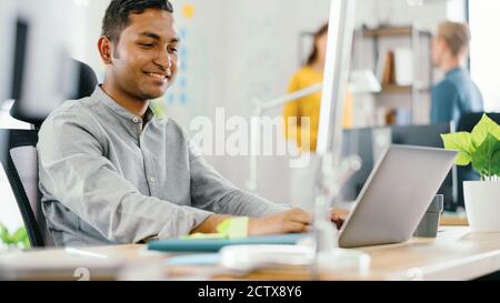 Gutaussehende lächelnde indische Büroangestellter sitzen an seinem Schreibtisch arbeitet auf einem Laptop. Im Hintergrund modernes Büro mit vielfältigen Team von Young Professionals Stockfoto