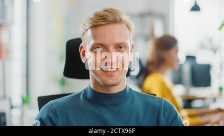 Porträt eines gutaussehenden professionellen Kaukasischen Mann, der an seinem Schreibtisch arbeitet und lächelnd auf die Kamera schaut. Erfolgreicher Mann, der in Bright Diverse Office arbeitet. Stockfoto