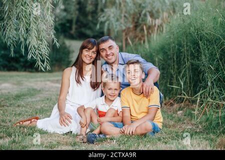 Schöne glückliche kaukasische Familie von vier Personen im Park am Sommertag. Mutter und Vater umarmen zwei Söhne Brüder Geschwister. Authentischer Lifestyle Stockfoto