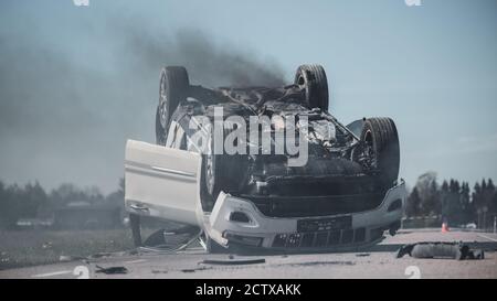 Horrific Verkehrsunfall Rollover Rauchen und brennendes Fahrzeug auf seinem Dach in der Mitte der Straße nach Kollision liegen. Crash-Szene tagsüber mit Stockfoto