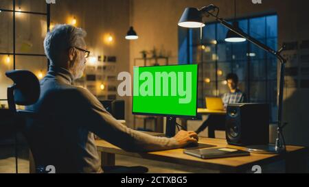 Kreativer Designer mittleren Alters, der an seinem Schreibtisch sitzt, verwendet Desktop-Computer mit grünem Mock-up-Bildschirm. Am Abend arbeiten zwei Creative-Mitarbeiter an Stockfoto