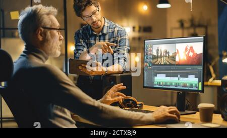 Abend in Creative Office: Professionelle Middle Aged Videographer arbeitet auf Desktop-Computer, hat Gespräch mit Younger Specialist, der Tablet verwendet Stockfoto