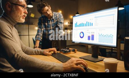 Professional Senior Business Martket Analyst arbeitet auf Desktop-Computer Arbeiten mit Charts, Grafiken, hat Gespräch mit Younger Specialist, der gibt Stockfoto