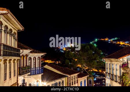 Fassaden von Häusern in Kolonialarchitektur auf einem alten Kopfsteinpflaster Straße in der Stadt Ouro Preto beleuchtet in der Nacht Mit historischer Kirche in backgro Stockfoto