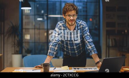 Junger kreativer Designer, der an seinem Schreibtisch arbeitet, Smiles on Camera. Er macht Papierkram, wählt das richtige Design, Konzept und Skizzen. Im Hintergrund Stockfoto