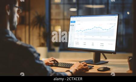 Über die Schulter selbstbewusster junger Geschäftsmann, der an seinem Schreibtisch sitzt und Desktop-Computer mit Statistiken, verschiedenen Daten, Diagrammen und Grafiken verwendet. Abend in Stockfoto