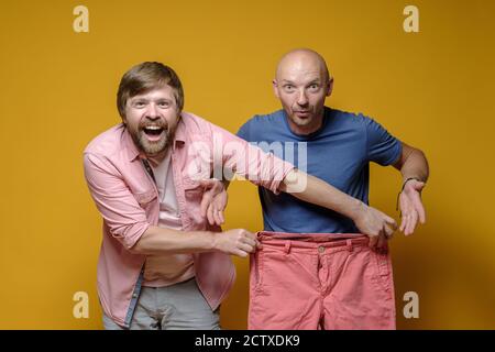 Zwei Freunde kaufen ein. Fröhlicher Mann versuchte scherzhaft auf große Hosen für einen anderen perplexen Männer. Verkaufskonzept. Gelber Hintergrund. Stockfoto