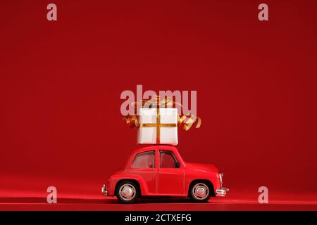 Rotes kleines Retro-Spielzeugauto mit Geschenk auf dem Dach auf rotem Hintergrund. Lieferung. Neujahr, Weihnachten, Valentinstag, Weltfrauentag, Verkaufskonzept. Stockfoto