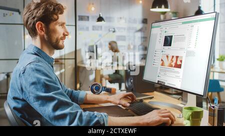 Schöne männliche Scrolls durch Social Media Site auf seinem Personal Computer mit großem Display. Er arbeitet in einem Cool Office Loft. Andere Weibliche Kreative Stockfoto