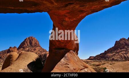 Natürlicher orangefarbener Felsbogen mit abgerundeten Felsen und Bergen im Hintergrund bei Spitzkoppe, Kalahari Wüste, Namibia, Afrika. Stockfoto