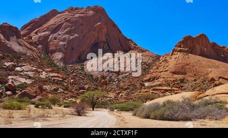 Vorderansicht der roten Spitzkoppe, Kalahari Wüste, Namibia, Afrika mit Schotterstraße und Vegeation in heißer Mittagssonne. Stockfoto