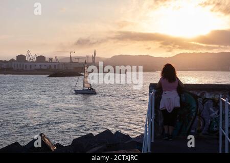 Sportliche Frau, die den Sonnenuntergang am Meer genießt. Segelboot, das in Richtung Hafen segelt, Kräne im Hintergrund. Graffiti an der Wand. Gijón, Asturien, N Stockfoto