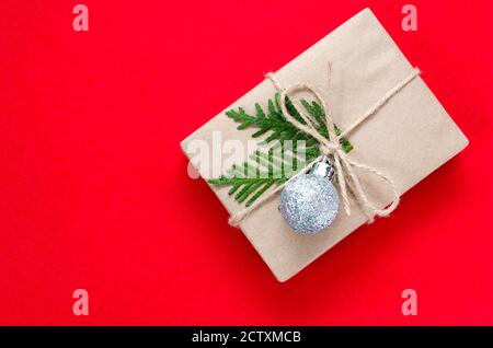 Geschenk- oder Geschenkbox in Kraftpapier verpackt, verziert mit silberner Weihnachtskugel und grünem Tannenzweig auf rotem Hintergrund. Urlaubskonzept. Flach liegend Stockfoto