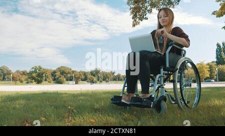 Remote-Work-Konzept. Junge schöne behinderte kaukasische Frau im Rollstuhl mit Laptop in der Natur. Hochwertige Fotos Stockfoto