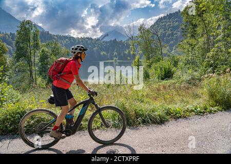 Nette ältere Frau mit ihrem Elektro-Mountainbike über dem Freiberger See und einer großen Skiflugschanze in den Allgauer Alpen bei Oberstdorf, Deutschland Stockfoto