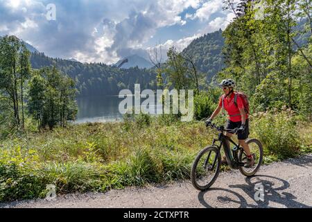 Nette ältere Frau mit ihrem Elektro-Mountainbike über dem Freiberger See und einer großen Skiflugschanze in den Allgauer Alpen bei Oberstdorf, Deutschland Stockfoto